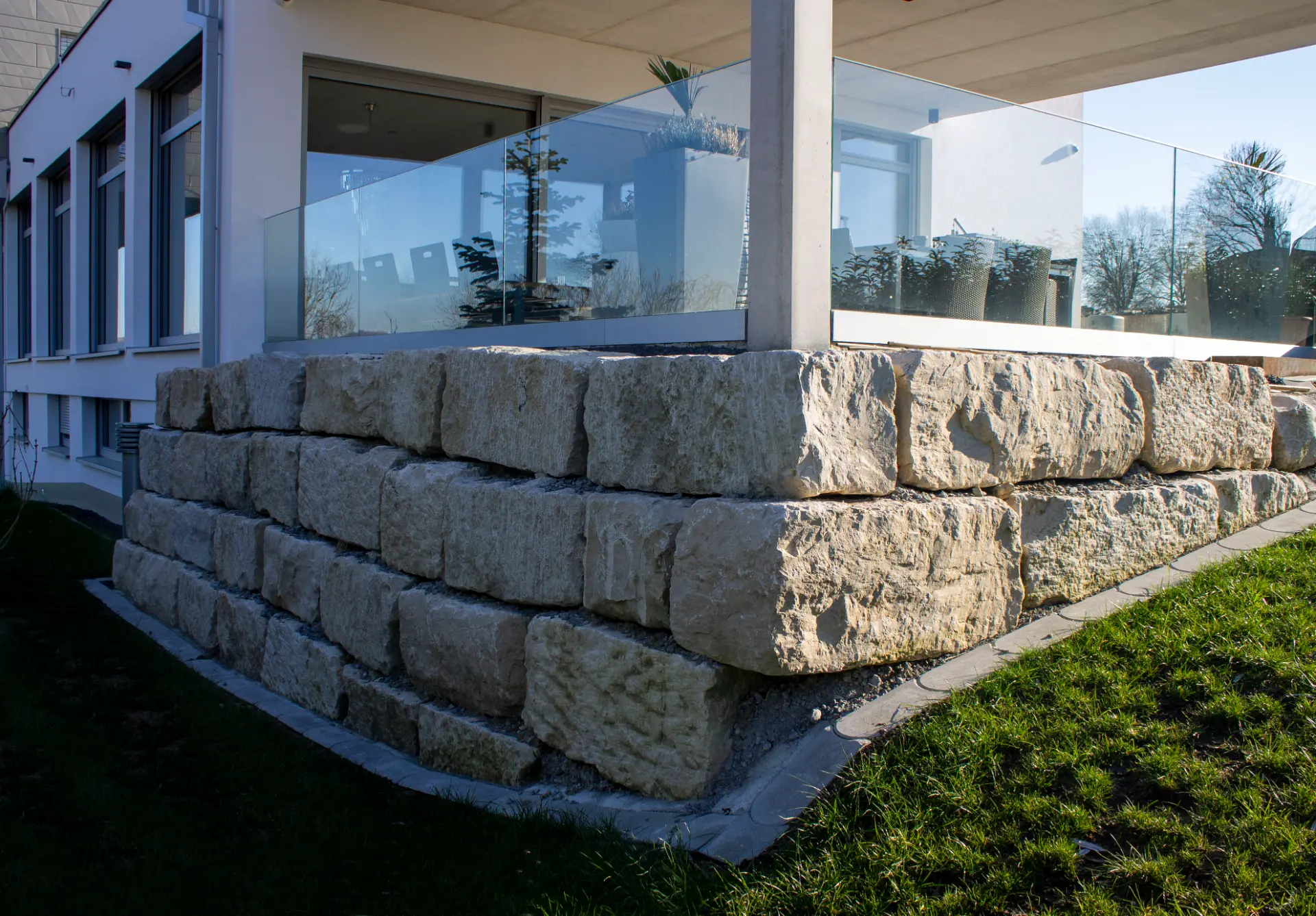 Stützmauer aus hellen Natursteinblöcken unter einer Terrasse mit Glasbrüstung