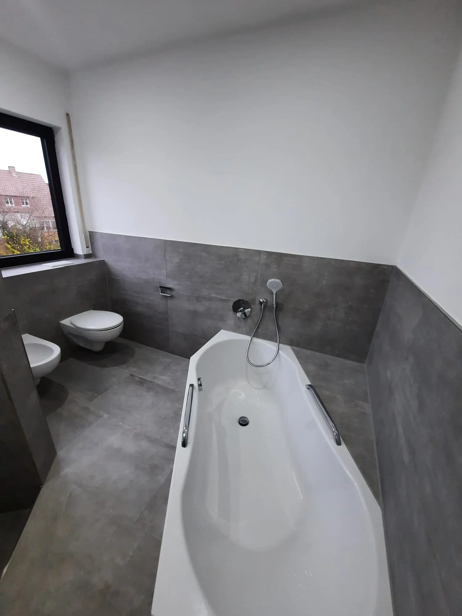 Fertiges Bad mit Einbauwanne, wandhängendem WC und Bidet vor grauen Großformatfliesen